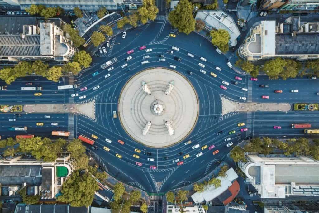 Aerial view of multi-lane traffic roundabout, with vehicles on main road running across left to right, surrounded by trees and buildings.