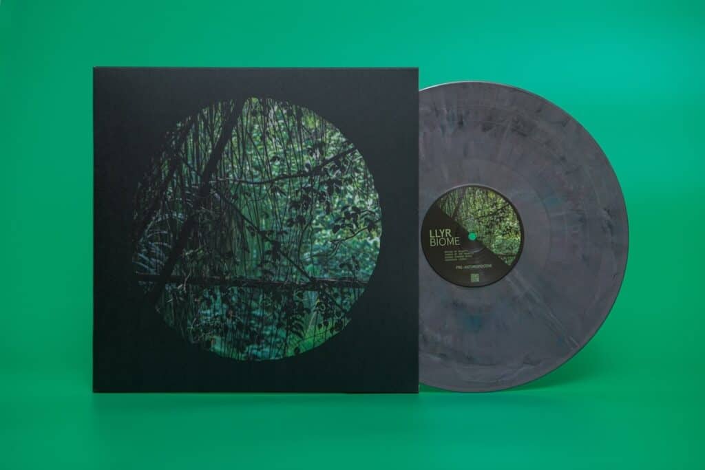 Variegated grey vinyl record mostly removed from sleeve featuring image of trees, with name Llyr Biome visible on label, against green background.