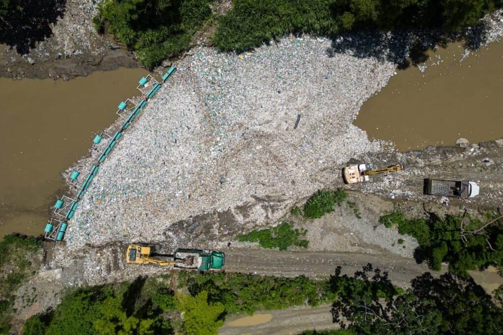 Aerial view of huge volume of trash trapped by interceptor in area of river, between banks, with excavator lifting loads onto roadside truck.