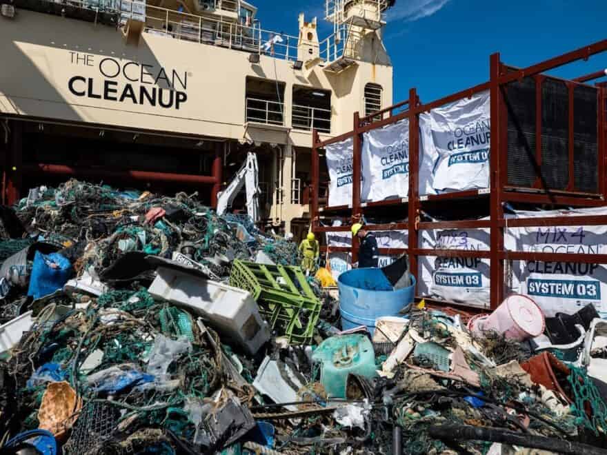 Mass of plastic debris on board ship, ready for sorting and storing, with Ocean Cleanup name visible across bridge of vessel above.