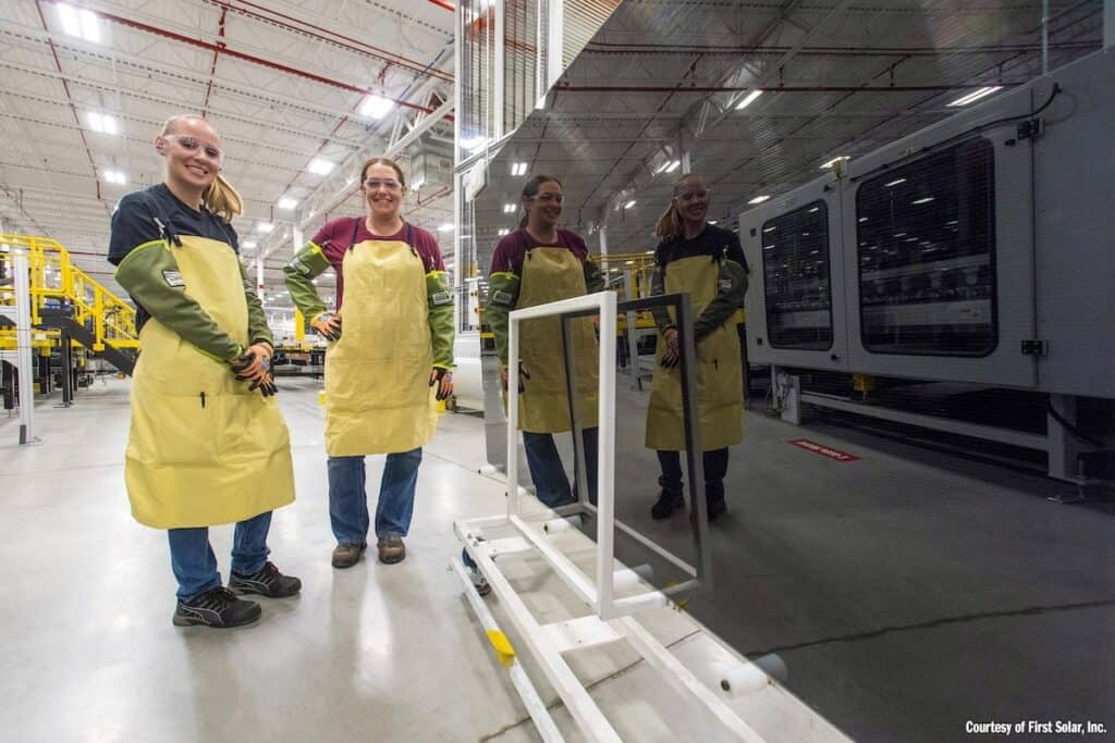 Two manufacturing personnel in yellow aprons stand beside a large solar PV module component, in which they and the factory are reflected.