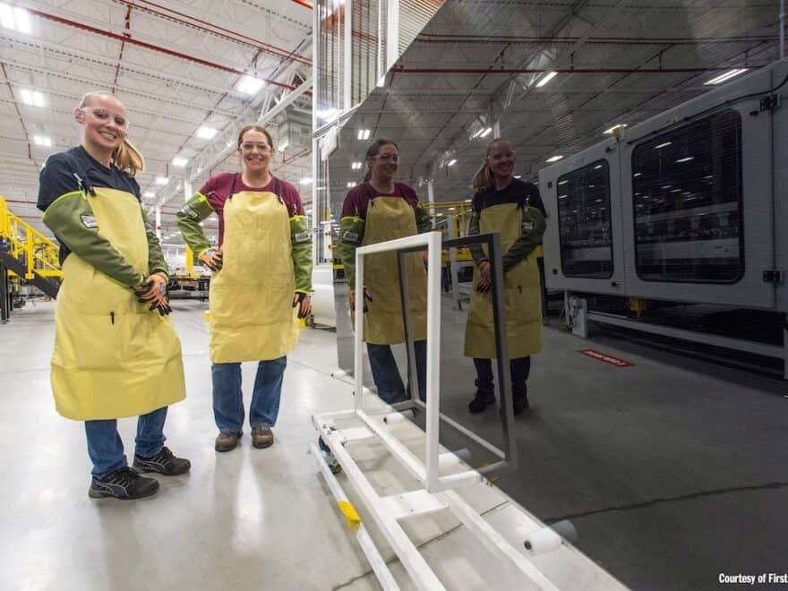 Two manufacturing personnel in yellow aprons stand beside a large solar PV module component, in which they and the factory are reflected.