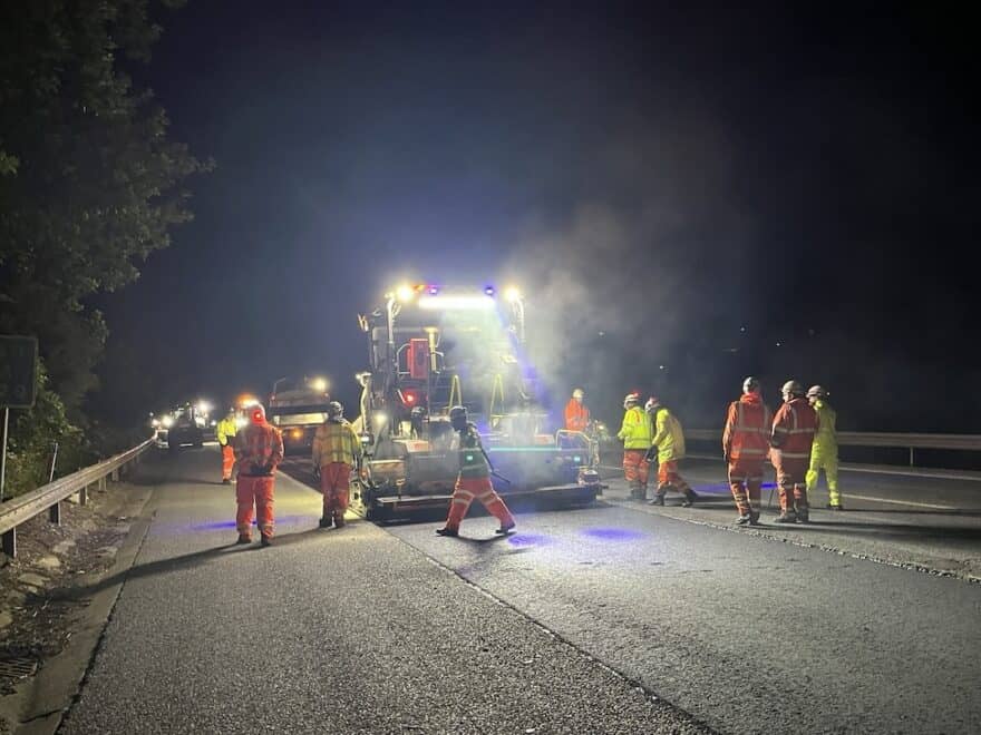 Roadworkers in orange and yellow high-vis clothing laying new surface on M11 motorway under lights of heavy machinery.