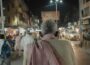 Back on elderly man's head as he walks through busy city street at night.