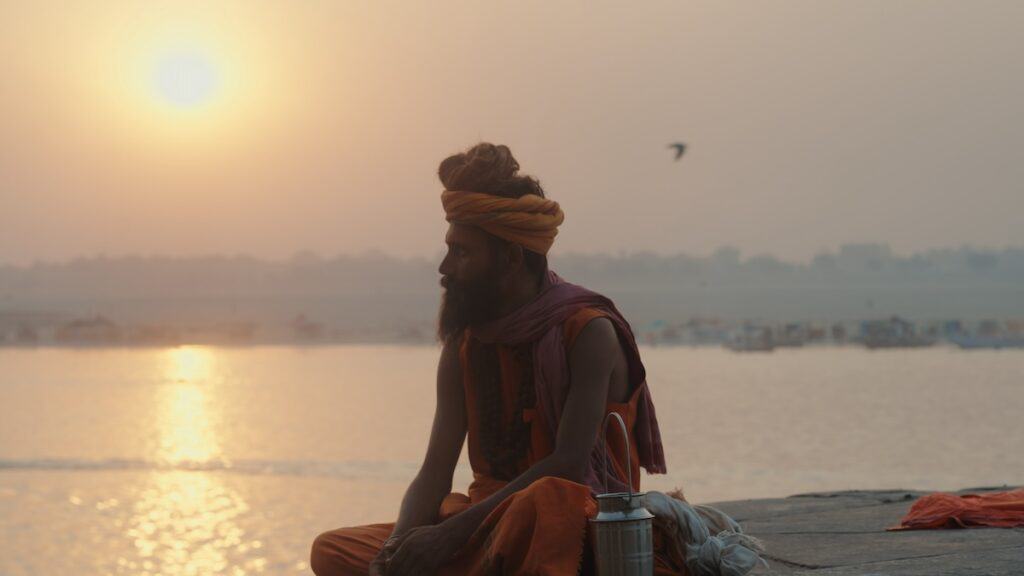 Bare-armed bearded man in traditional orange robes and turban by waters of Ganges River at sunset.