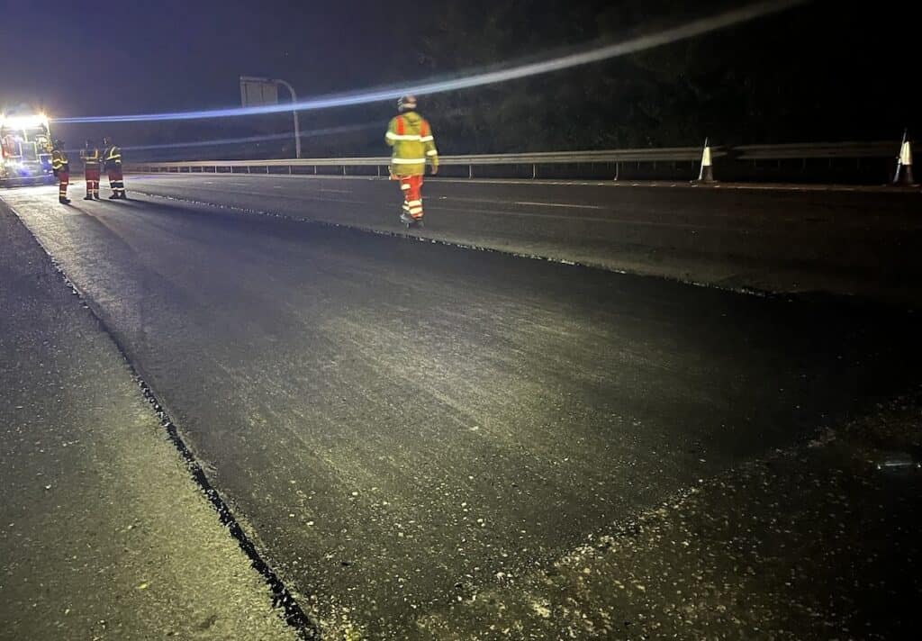 Roadworkers in orange and yellow high-vis clothing beside new surface on M11 motorway, with lights of heavy machinery in background.