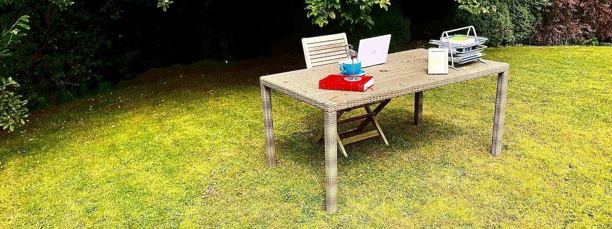 Desk in outdoor garden setting, complete with laptop, in-trays, books, framed photo, ornament, cup and saucer, plus with trees behind.