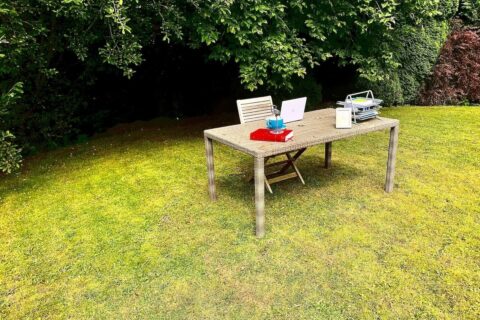 Desk in outdoor garden setting, complete with laptop, in-trays, books, framed photo, ornament, cup and saucer, plus with trees behind.