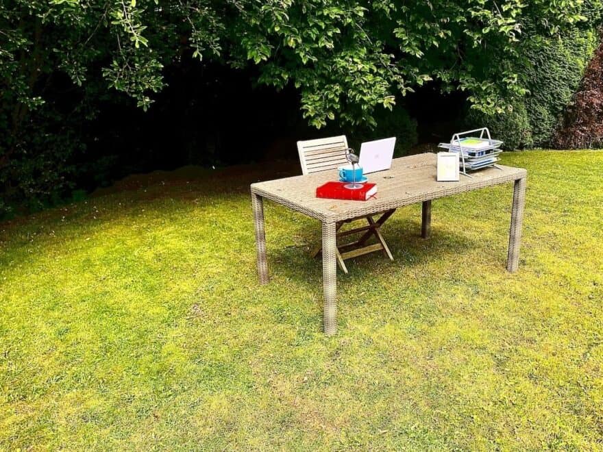 Desk in outdoor garden setting, complete with laptop, in-trays, books, framed photo, ornament, cup and saucer, plus with trees behind.