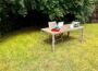 Desk in outdoor garden setting, complete with laptop, in-trays, books, framed photo, ornament, cup and saucer, plus with trees behind.