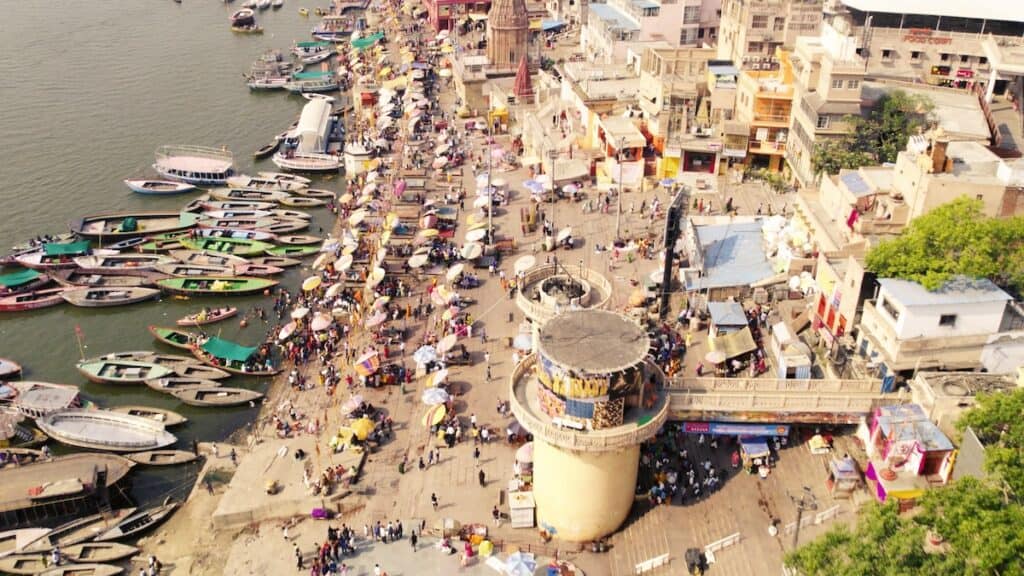 Aerial view of Varanasi waterfront, with row boats moored on Ganges River to left and sun umbrellas on beach.