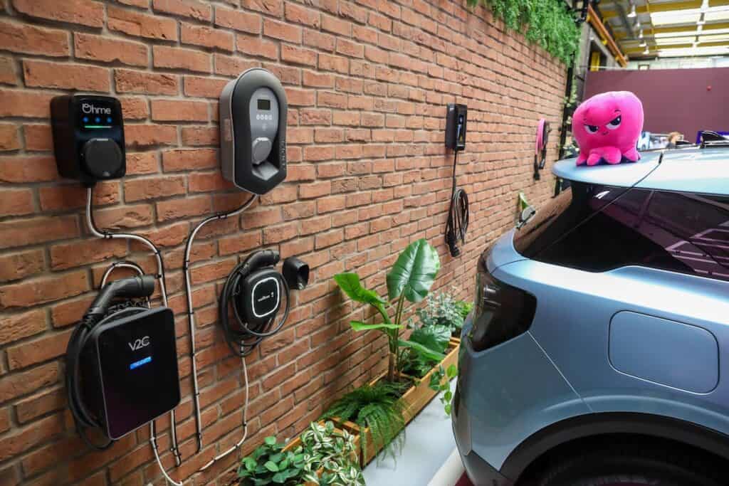 Rear of electric vehicle parked (with pink Octopus brand soft toy on roof) beside brick wall of EV charging tech, with green plants at base.