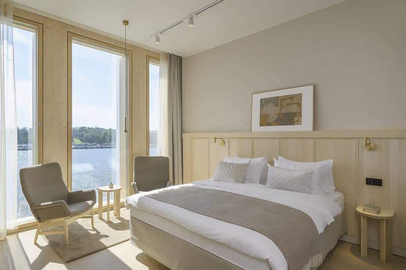 Hotel double bedroom in light wood and muted colours, with pair of modern brown upholstery armchairs beside full-height sea-view windows.