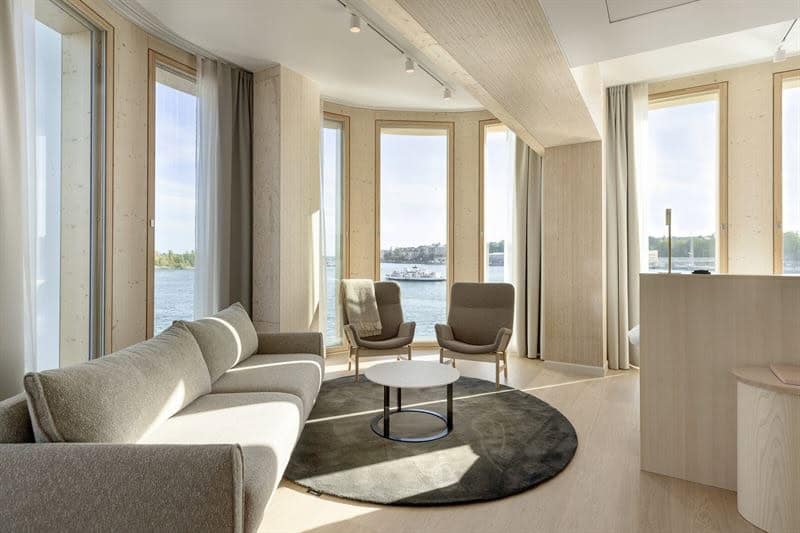 Lounge area of hotel suite in light wood and shades of brown, with settee, armchairs and coffee table, plus full-height sea-view windows.