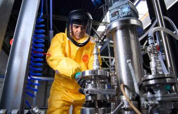 Person in bright yellow HAZMAT suit, complete with face mask, works on pilot test manufacturing equipment.