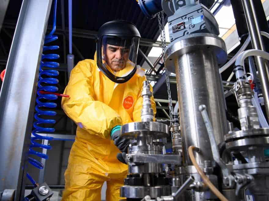 Person in bright yellow HAZMAT suit, complete with face mask, works on pilot test manufacturing equipment.