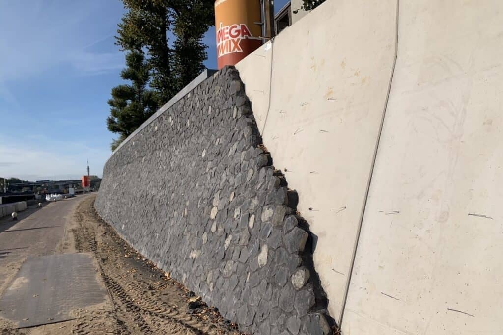 Construction of retaining wall showing precast concrete beneath finish of reused basalt stones, plus orange MegaMix concrete units visible above.