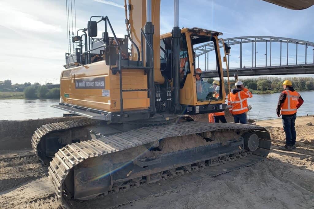 Yellow construction plant digger with caterpillar tyres by river, plus workers in orange high-vis and hard hats alongside.