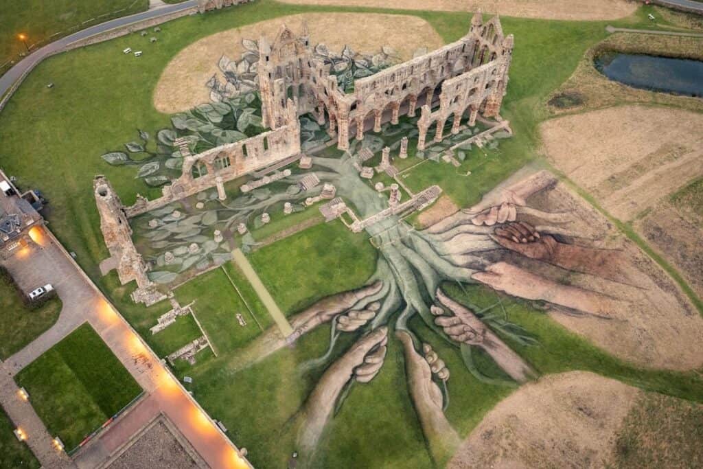 Aerial view of land art installation in and around ruins of Whitby Abbey, with hands drawn holding roots of tree in natural earth greens and browns.