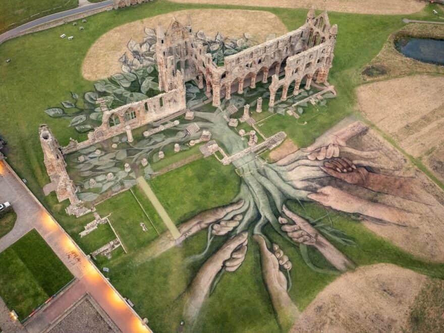 Aerial view of land art installation in and around ruins of Whitby Abbey, with hands drawn holding roots of tree in natural earth greens and browns.