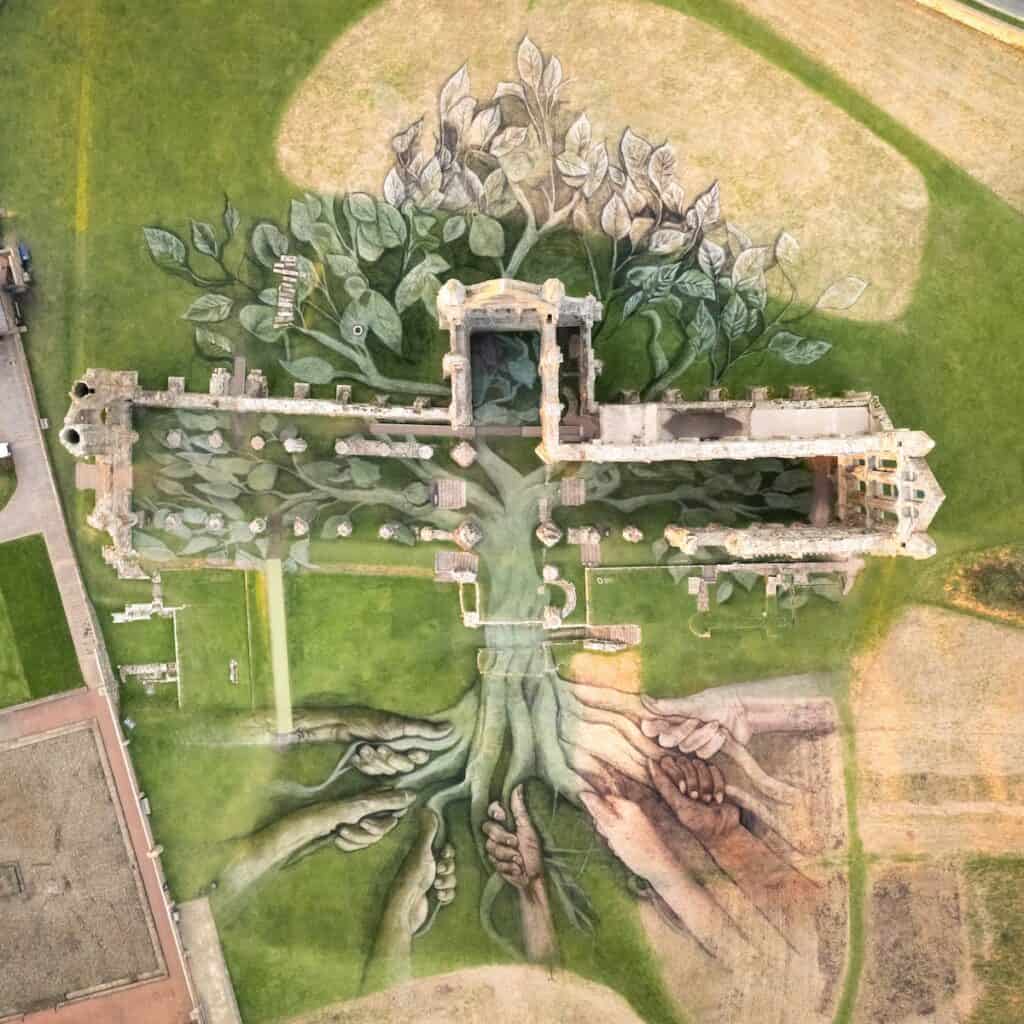 Full aerial view of land art installation in and around ruins of Whitby Abbey, with hands drawn holding roots of tree in natural earth greens and browns.