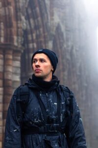 Portrait of artist David Popa, in blue woollen hat and waterproof coat, spattered with white paint, against misty backdrop of ruined Abbey walls.