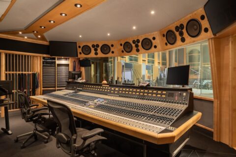 Recording studio mixing desk with two chairs and huge multi-track console, plus speakers above window looking out into performance space.