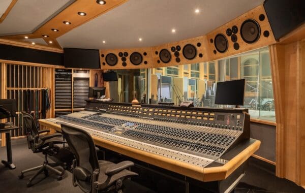 Recording studio mixing desk with two chairs and huge multi-track console, plus speakers above window looking out into performance space.