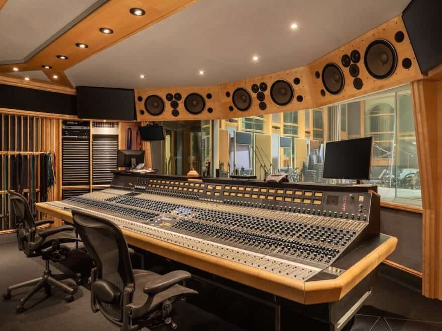 Recording studio mixing desk with two chairs and huge multi-track console, plus speakers above window looking out into performance space.