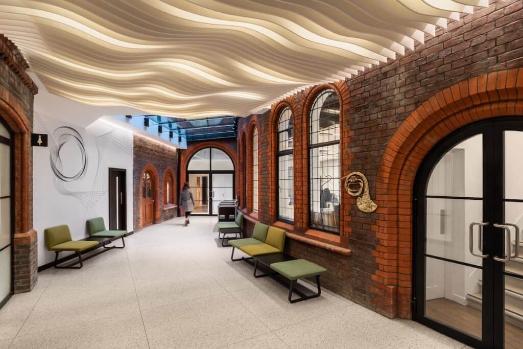 Internal corridor with green seating against brick exterior-like walls, arched windows and doorways, plus person exiting at far end.