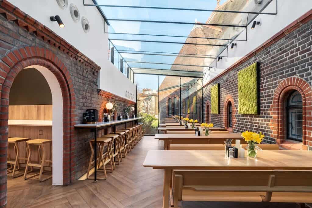 Internal café and bar area, with brick exterior-like walls, wooden bench tables and seating along wall, plus glazed ceiling overhead.