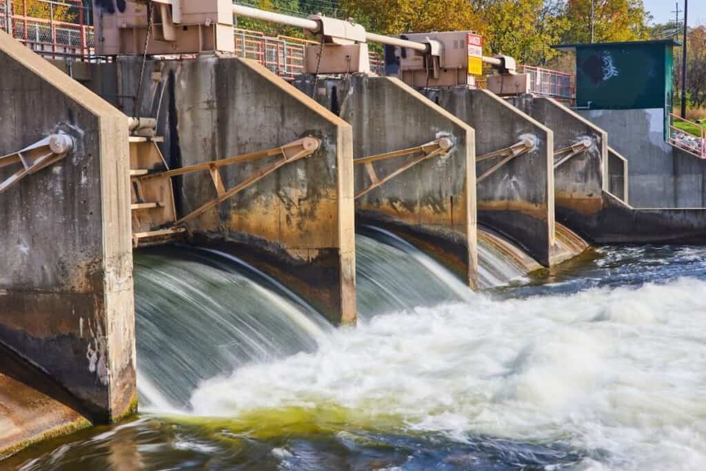 Close-up of water surging through top of concrete dam structure, foaming white, with blue beyond and some blue-green areas to foreground.