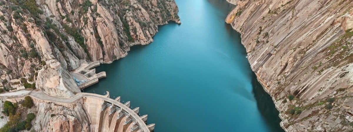 Aerial shot showing dam in bottom left holding back lake of azure blue water, in deep mountain valley, with bare rock both sides.