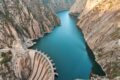 Aerial shot showing dam in bottom left holding back lake of azure blue water, in deep mountain valley, with bare rock both sides.