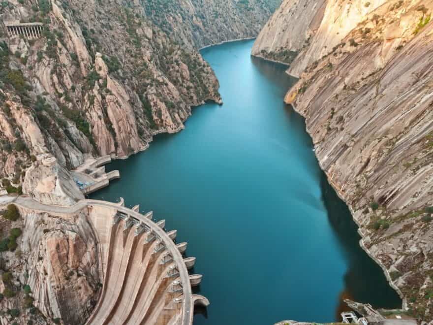 Aerial shot showing dam in bottom left holding back lake of azure blue water, in deep mountain valley, with bare rock both sides.