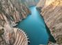 Aerial shot showing dam in bottom left holding back lake of azure blue water, in deep mountain valley, with bare rock both sides.