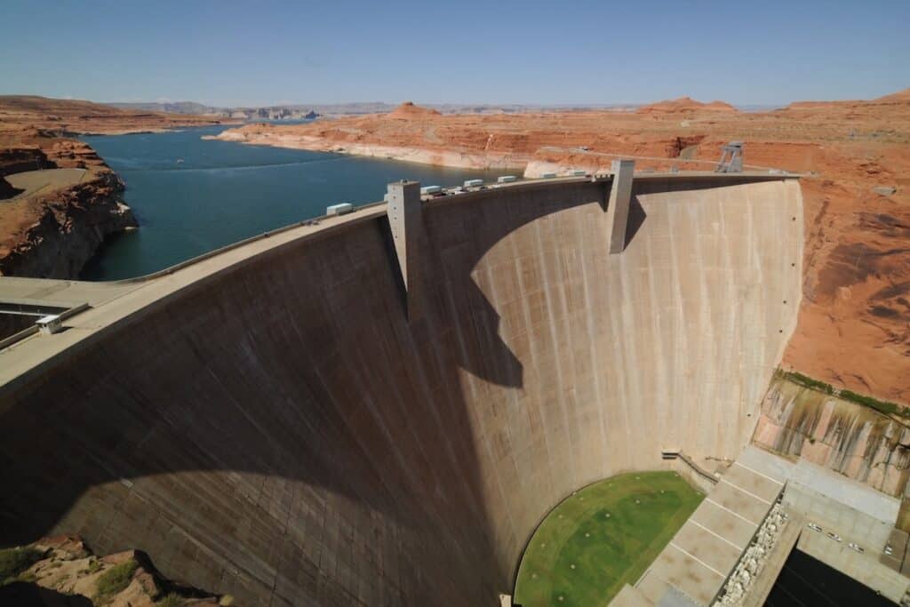 Aerial shot of dam up diagonally l-r, with deep blue river to left, grass-green semi-circle at base to right, all within red rock canyon both sides.