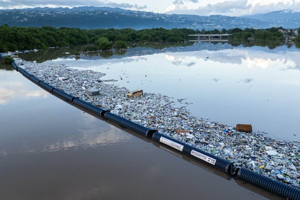 Aerial view of Interceptor Barrier 015 deployed straight across water in Sandy Gully, Jamaica; trash trapped behind, trees and hills in distance.