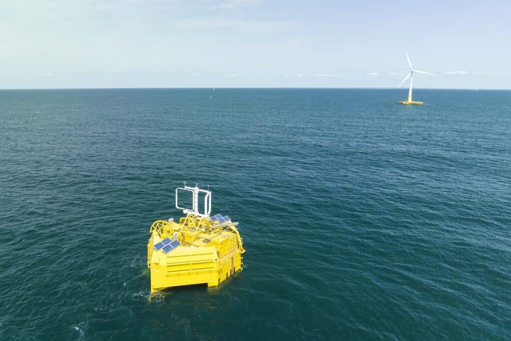 Bright yellow hydrogen production unit in foreground on left, with floating wind turbine on testbed in distance on right, both pictured out at sea.