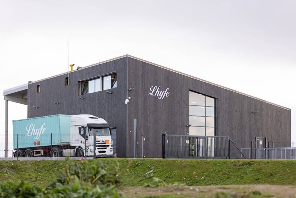 Truck carrying light blue container with Lhyfe branding in white leaves modern grey production facility, also with company name on side.