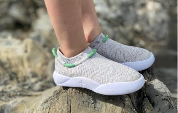 Side-on shot of Dubs Flex footwear in Dove Grey, with bare legs of wearer visible standing in rough stony landscape.
