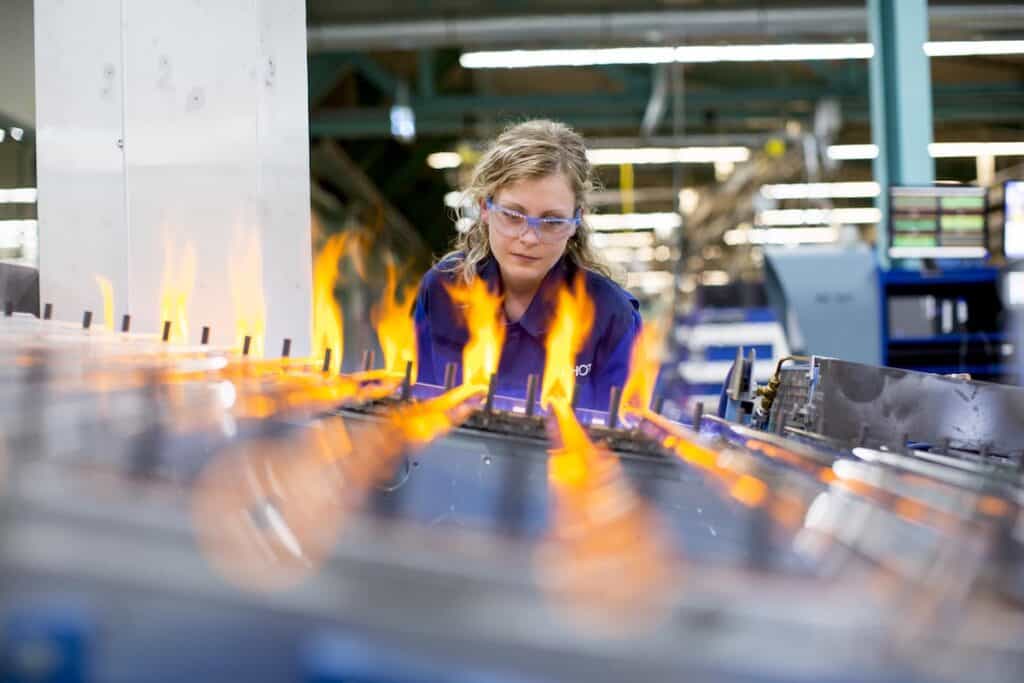 Female member of staff in branded Schott workwear and protective glasses oversees production on machine with flaming hot surface.