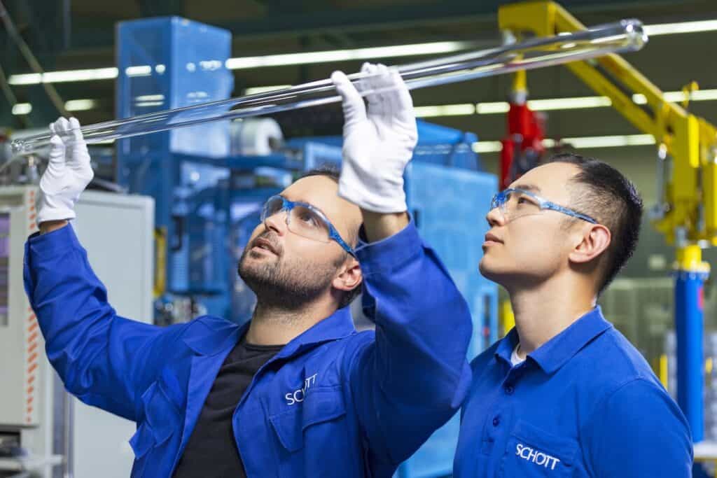 Two production staff members in blue Schott branded workwear and sporting protective eyewear, hold up and inspect long glass tube.