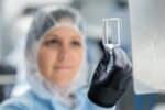 Out-of-focus female operative with hair covered in clear protective workwear holds small glass bottle clearly up to camera in gloved hand.