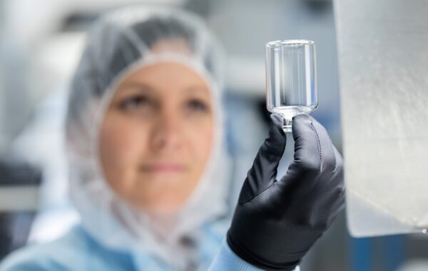 Out-of-focus female operative with hair covered in clear protective workwear holds small glass bottle clearly up to camera in gloved hand.