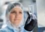 Out-of-focus female operative with hair covered in clear protective workwear holds small glass bottle clearly up to camera in gloved hand.