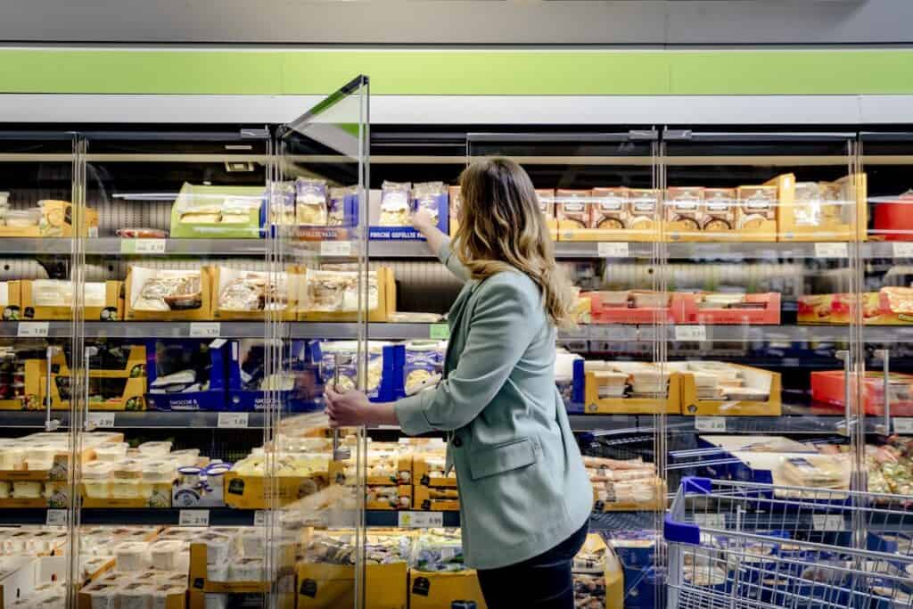 Long-haired female shopper with trolley cart opens glass door to chiller cabinet in food retail environment.