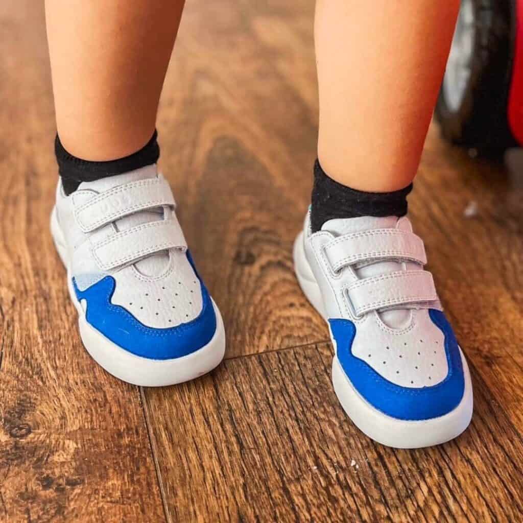 Glide Dubs footwear in blue, pictured on wood floor, with bare lower legs of young wearer shown in short black socks.