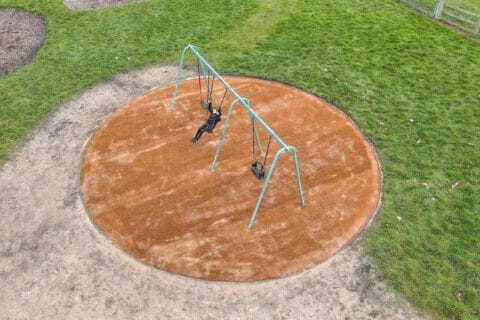 Aerial view of orangey-brown circle of CocoTurf play area in park, with young person on the swings, plus green grass and sandy areas beyond.