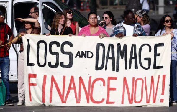 Diverse group of protestors stand behind homemade banner in white that says 'LOSS AND DAMAGE' in black above 'FINANCE NOW!' in red.